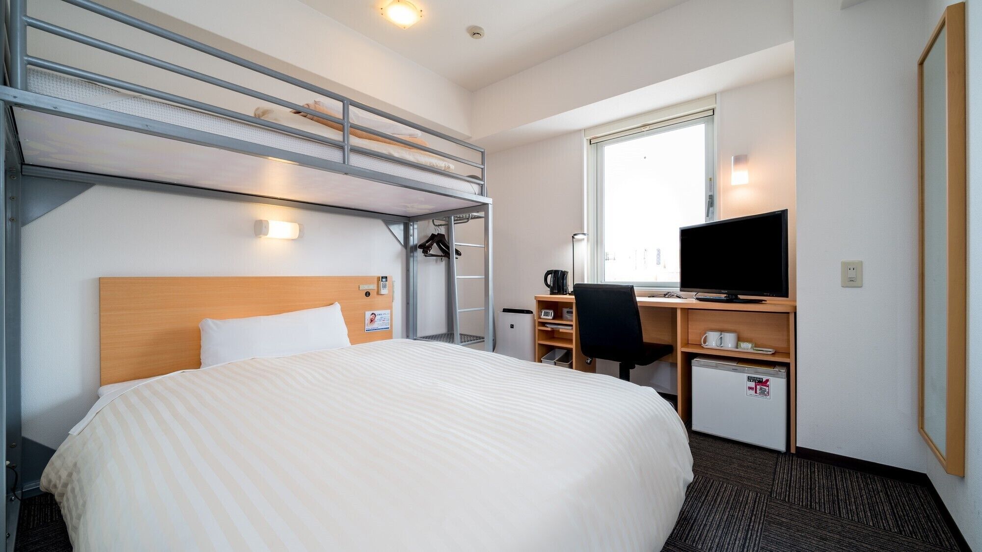 A hotel room with a bunk bed, a desk with a computer, a chair, a TV, and a small fridge.
