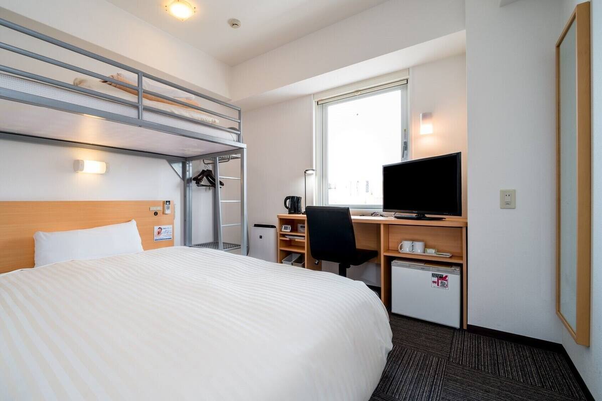 Habitación de hotel con literas, un escritorio con computadora, una silla, televisión y un refrigerador pequeño.
