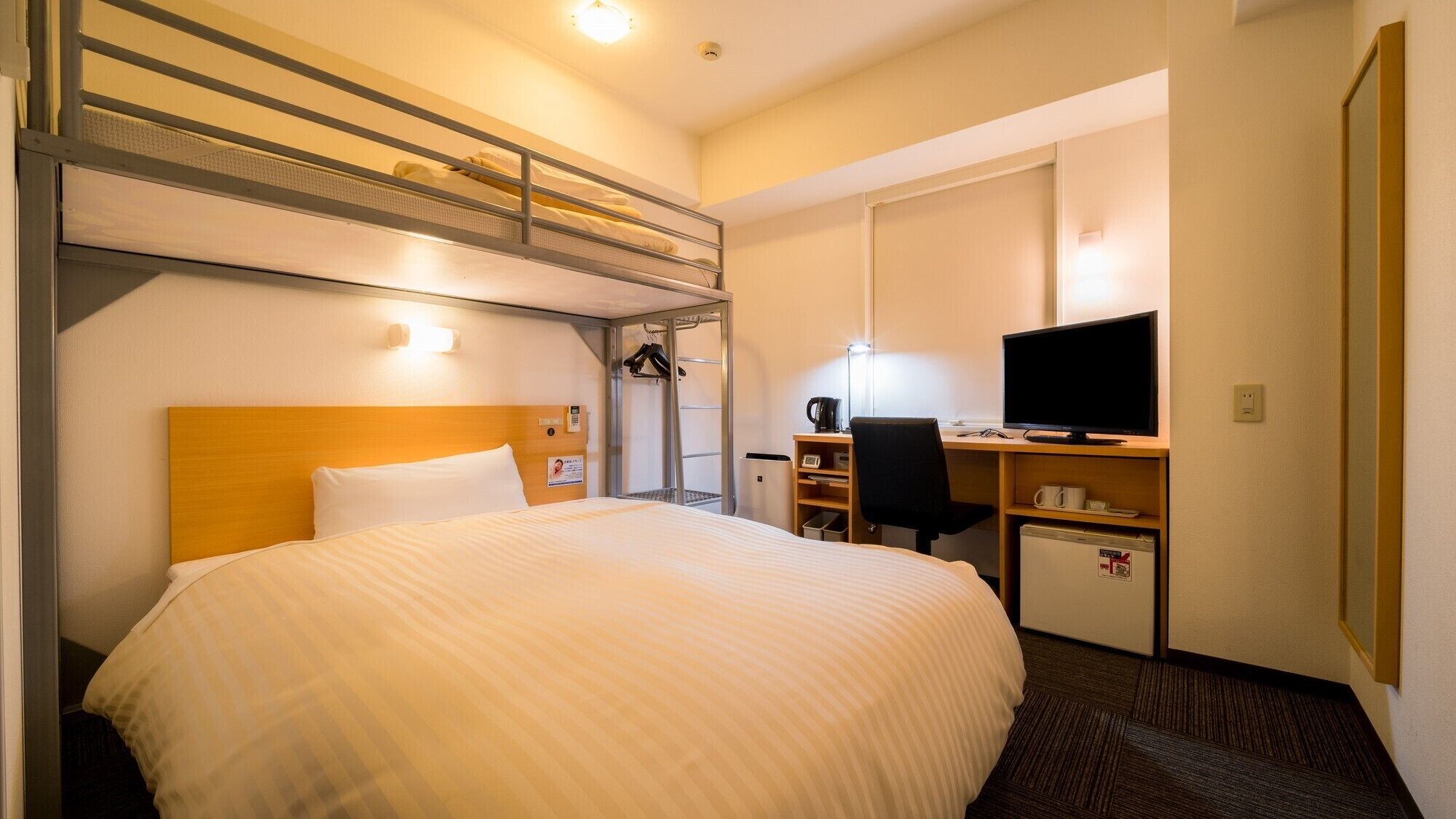 Een hotelkamer met een stapelbed, een bureau met een computer, een stoel en een kleine koelkast.