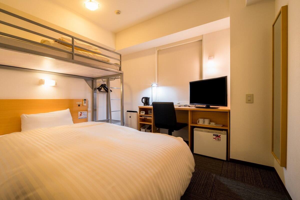 Una habitación de hotel con una cama litera, un escritorio con computadora, una silla y un refrigerador pequeño.