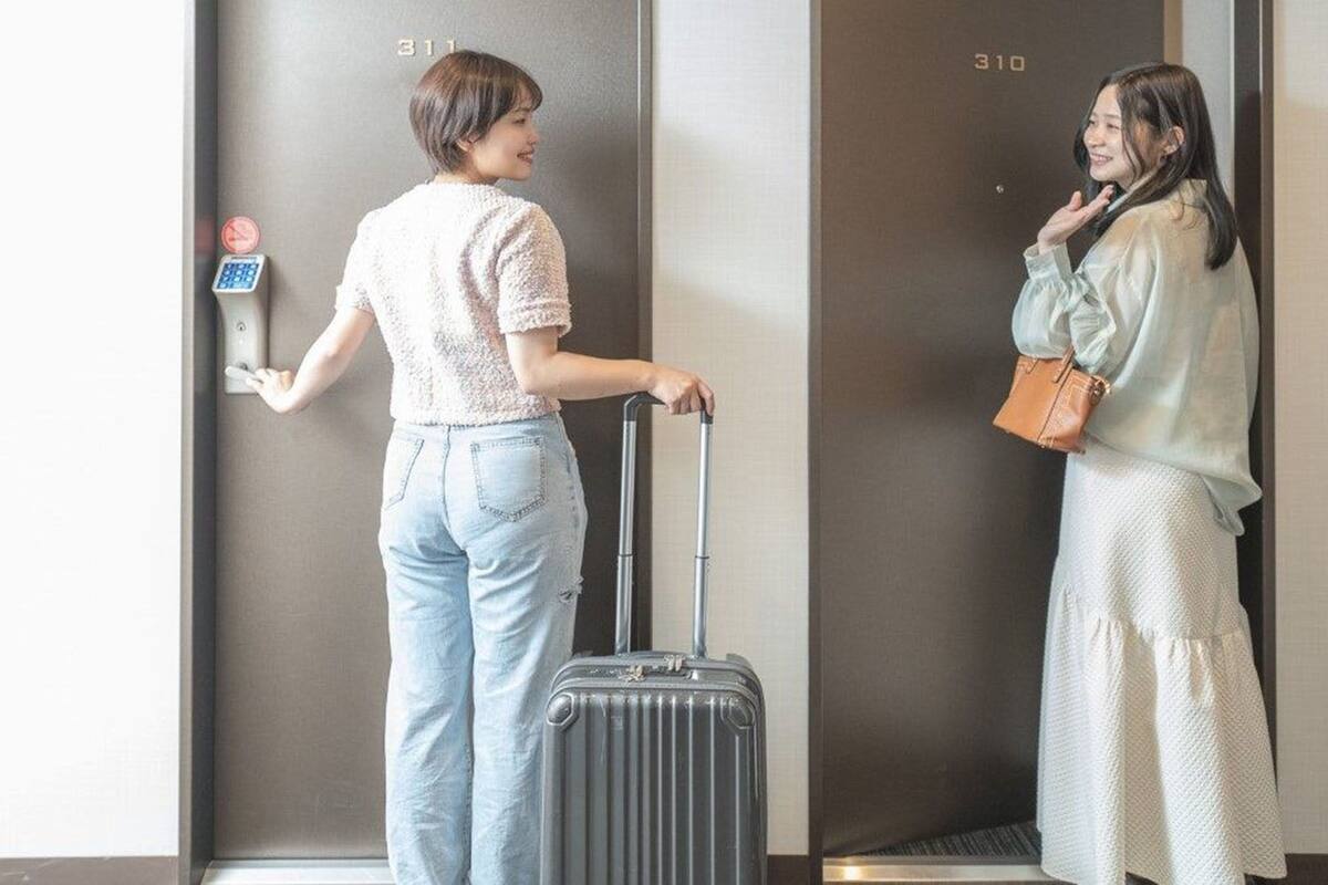 Dos personas están de pie en un pasillo con una puerta que indica '311' y '310'. Una de ellas lleva una maleta y un teléfono móvil.