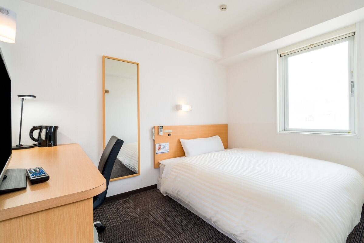 Una habitación de hotel con una cama, un escritorio con lámpara, un espejo y una ventana.