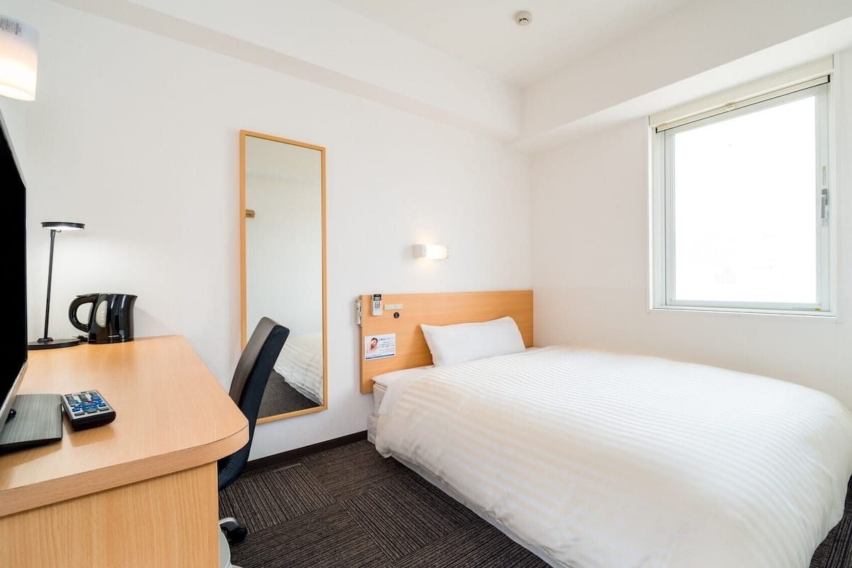 Une chambre d’hôtel avec un lit, un bureau avec une lampe, un miroir et une fenêtre.