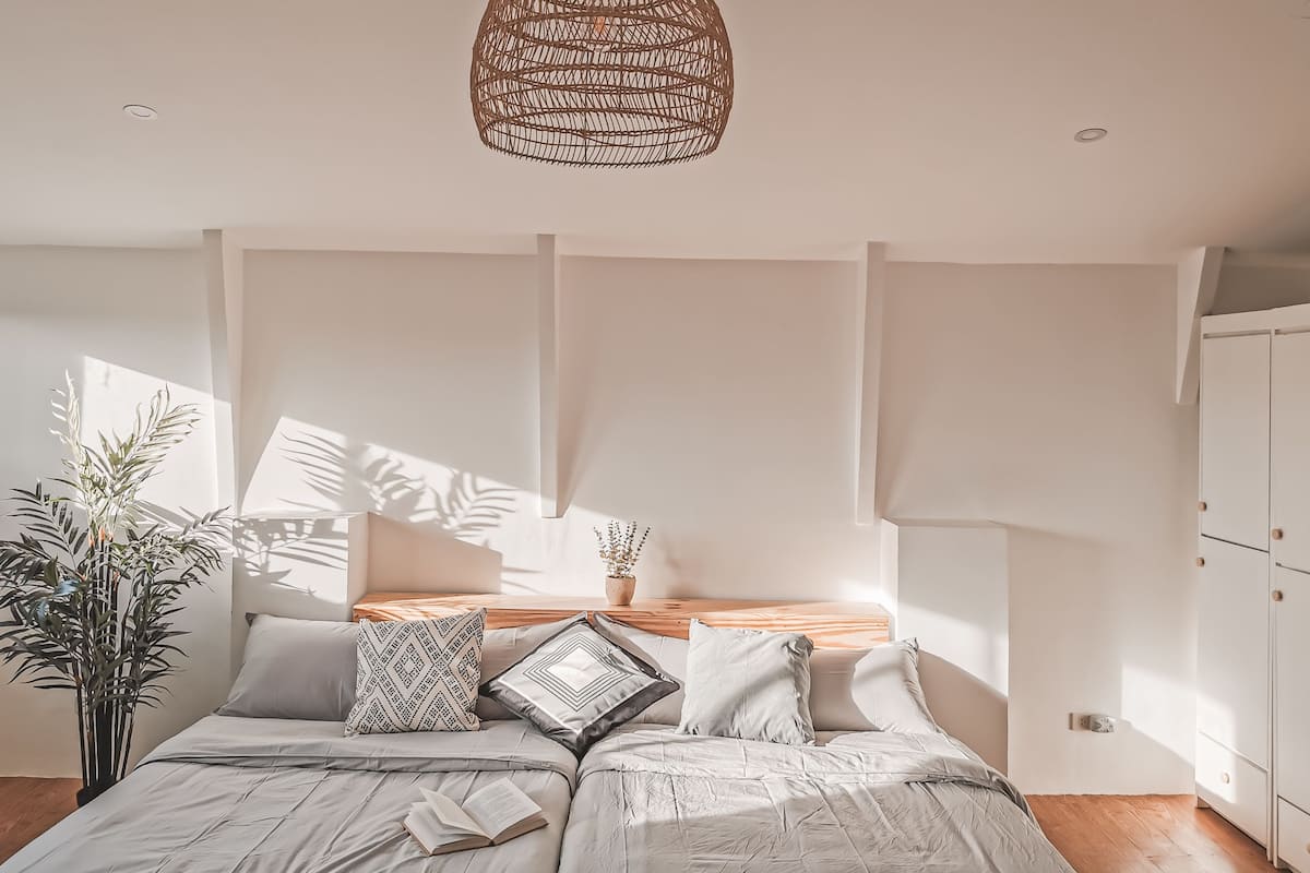 Une chambre à coucher avec un lit, des oreillers, un luminaire suspendu, une table de chevet et une plante en pot.