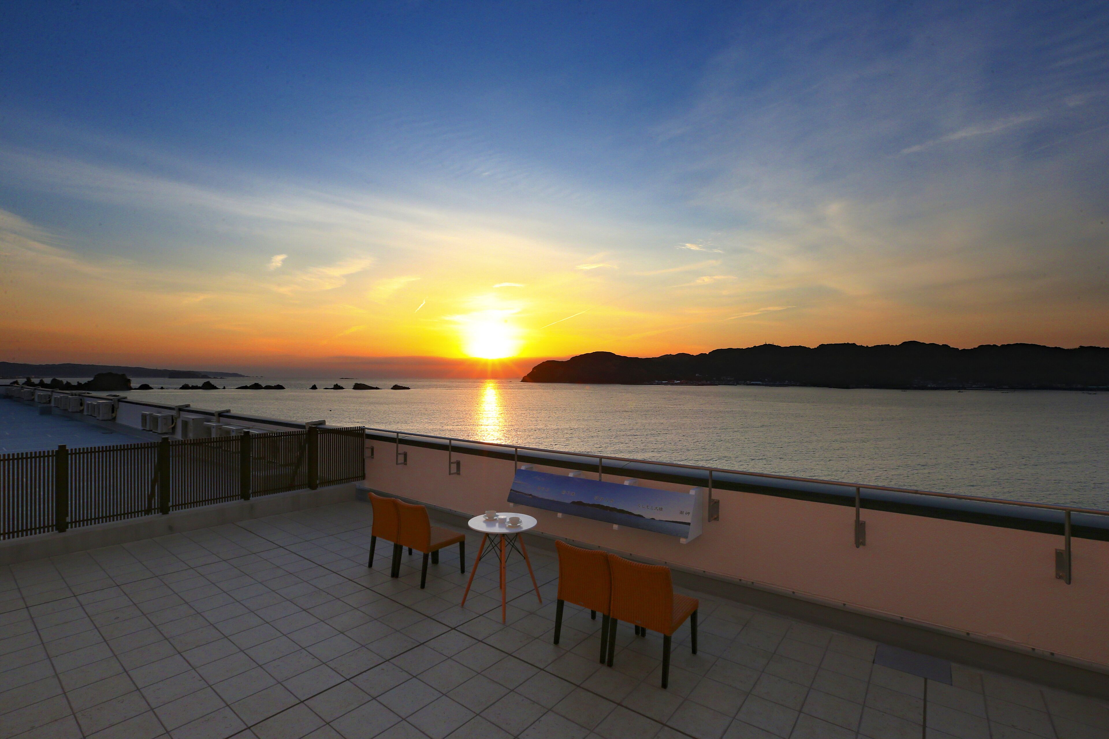 A terrace with two chairs and a table, overlooking a sunset over a body of water with distant landforms.
