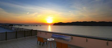 A terrace with two chairs and a table, overlooking a sunset over a body of water with distant landforms.