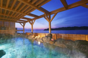 Zona de aguas termales con vigas de madera y rocas, con vista a un cuerpo de agua al anochecer.
