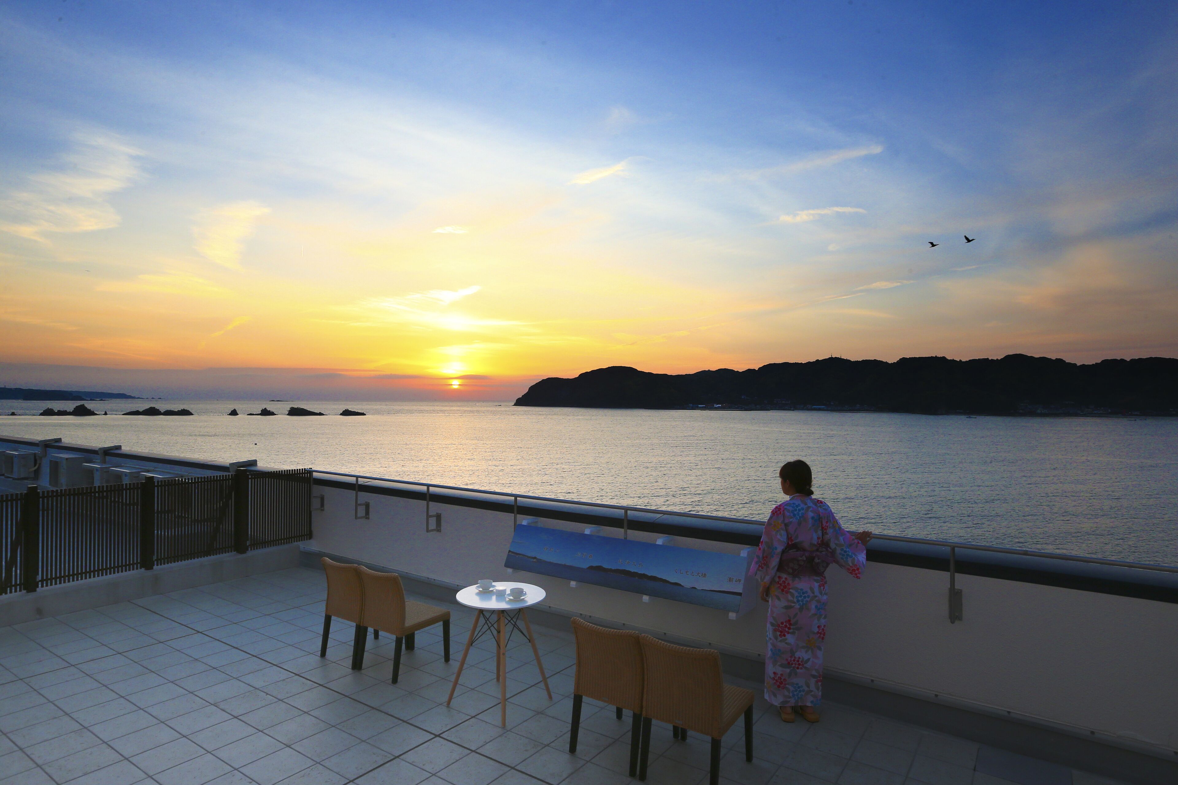 A balcony with a table and chairs, overlooking a body of water at sunset.