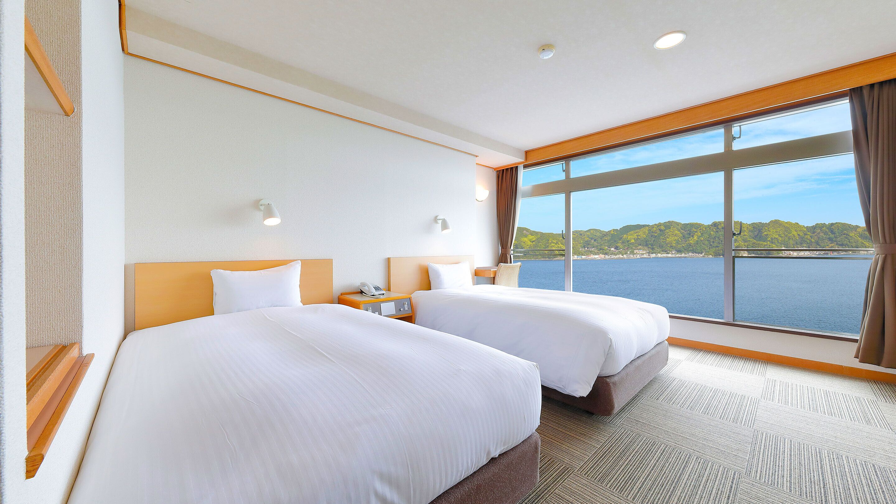 Ein Hotelzimmer mit zwei Betten, einem Nachttisch, einem Telefon und einem großen Fenster mit Blick auf Wasser und Grünflächen.