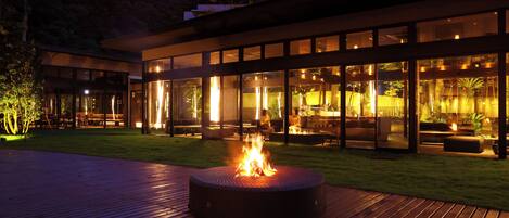 A modern building with large glass windows, a fire pit in the center, and outdoor seating areas.