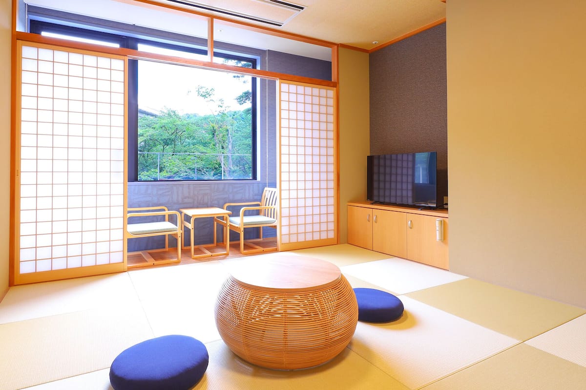 Une pièce de style japonais traditionnel, avec un sol recouvert de tatamis, une basse table en bois accompagnée de deux chaises, un téléviseur posé sur un meuble, et une vue sur la verdure à l’extérieur.