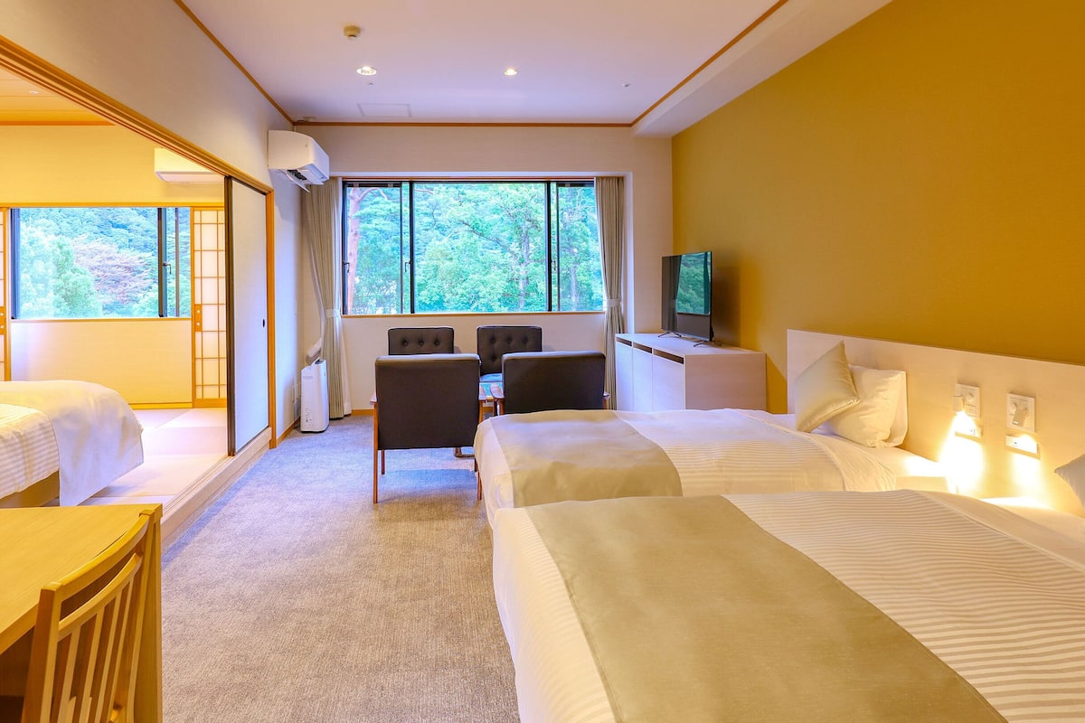 Une chambre d’hôtel avec deux lits, un bureau, une chaise, une télévision et de grandes fenêtres donnant sur la verdure.