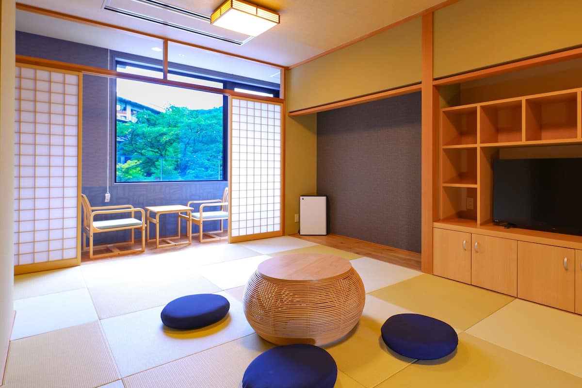 Une pièce de style japonais traditionnel, avec un sol recouvert de tatamis, une table basse, des coussins, un téléviseur et une vue sur la verdure à l’extérieur.