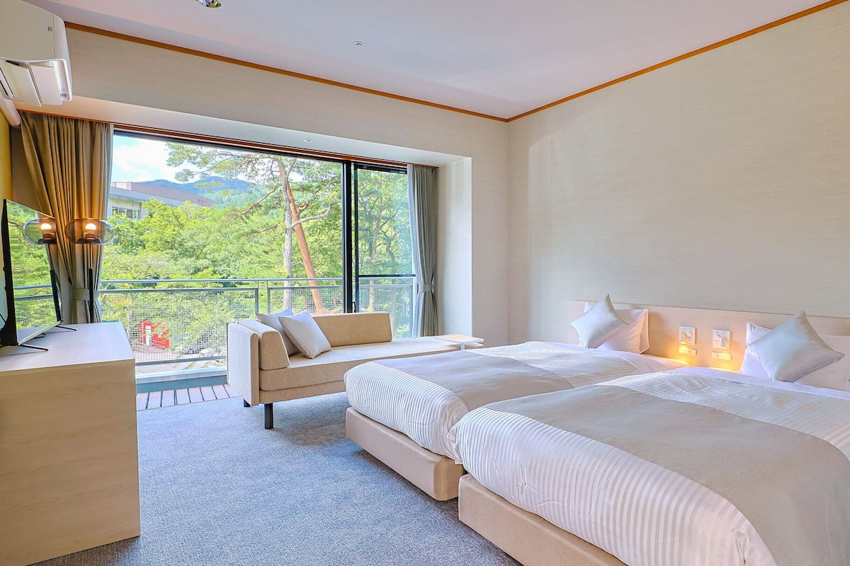 Une chambre d’hôtel avec deux lits, un canapé, un bureau et une grande fenêtre donnant sur un paysage verdoyant.