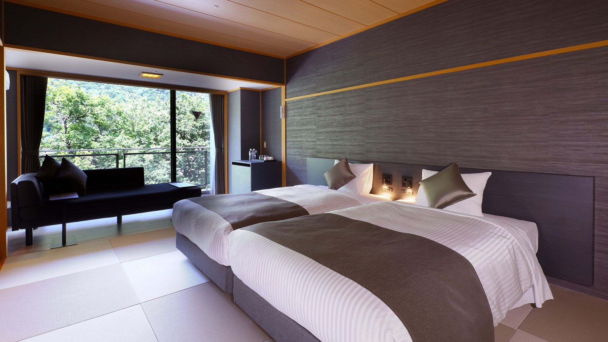 Une chambre d’hôtel moderne avec un grand lit, un coin salon près de la fenêtre et une vue sur la verdure environnante.