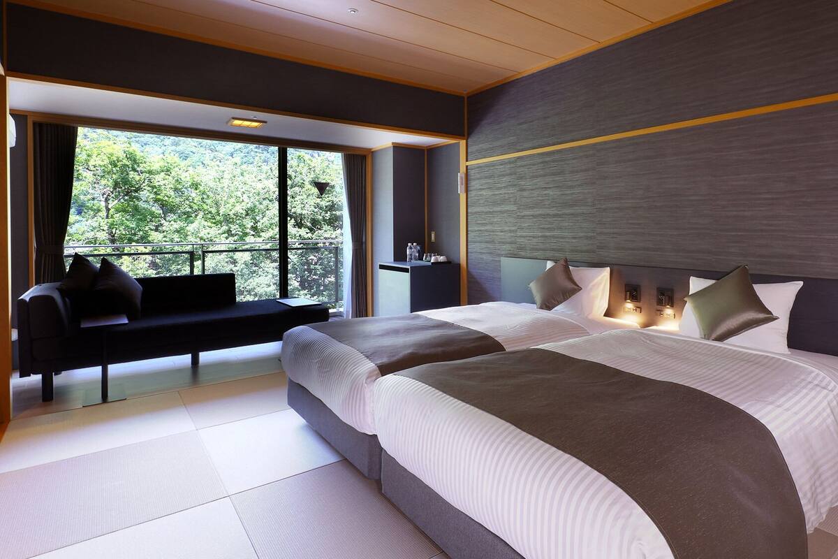 Une chambre d’hôtel moderne avec un grand lit, un coin salon près de la fenêtre et une vue sur la verdure environnante.