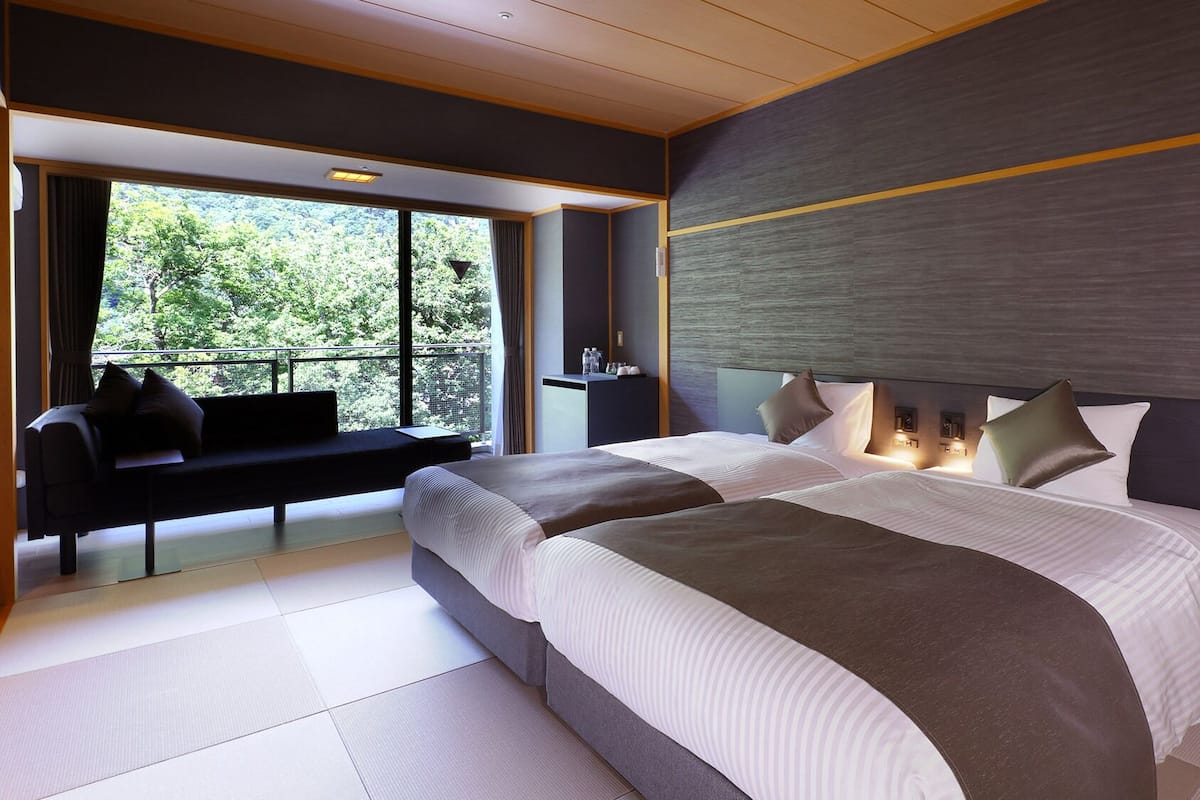 A modern hotel room with a large bed, a seating area by the window, and a view of greenery outside.