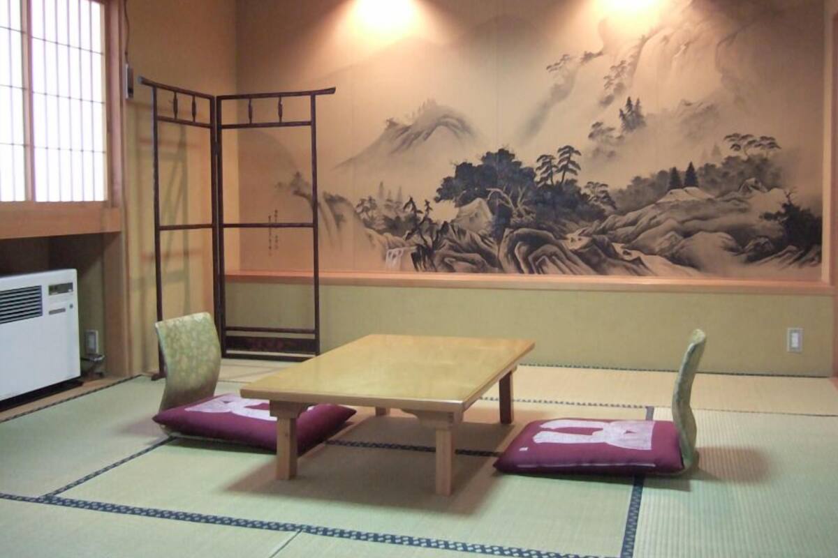 A traditional Japanese room with tatami flooring, a low wooden table, two cushions, a folding screen with a mountain landscape painting, and a standing heater.
