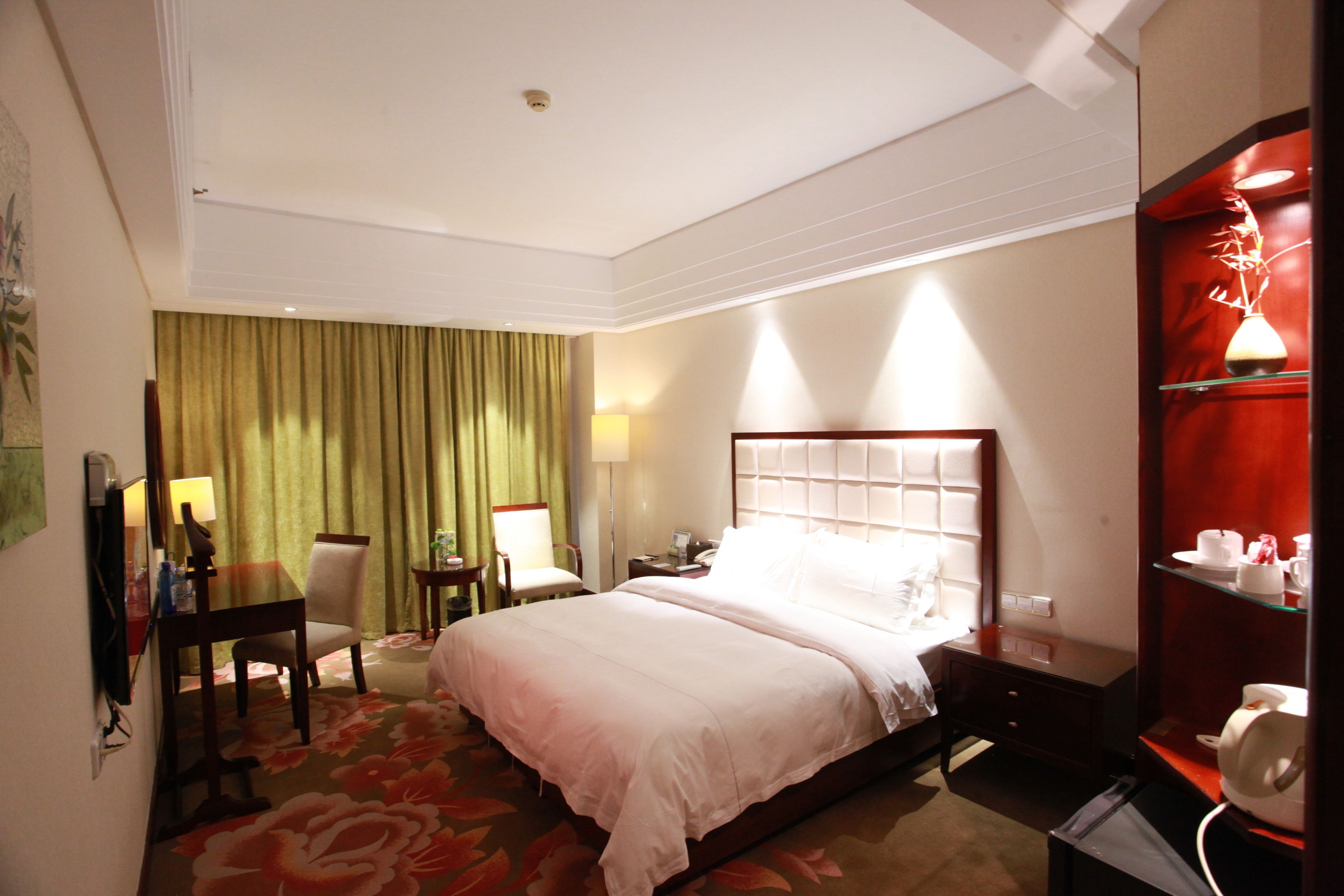 A hotel room with a bed, a desk, a chair, a lamp, and a shelf with cups and saucers.