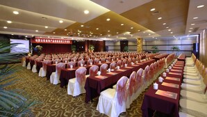 A conference room with rows of tables and chairs, a stage with a red backdrop, and a large screen.