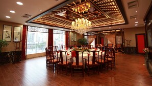 A spacious dining area with a high ceiling, chandeliers, and a large window offering a city view.
