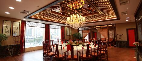A spacious dining area with a high ceiling, chandeliers, and a large window offering a city view.