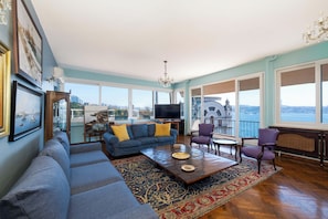 A spacious living room with a large sofa, a coffee table, and a television. There are large windows offering a view of the city. - Waterfront Penthouse Apt. with a Stunning View (İstanbul)