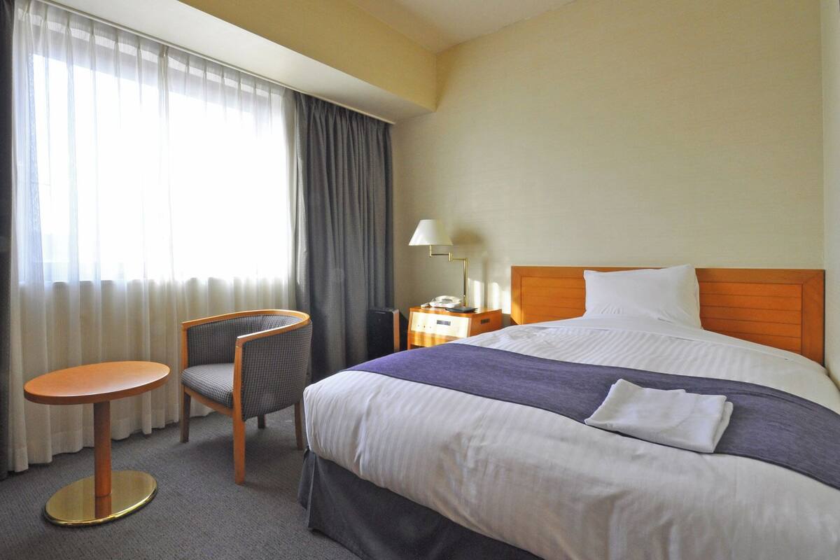 Quarto de hotel com cama, uma cadeira, uma mesinha, mesa de cabeceira e abajur.