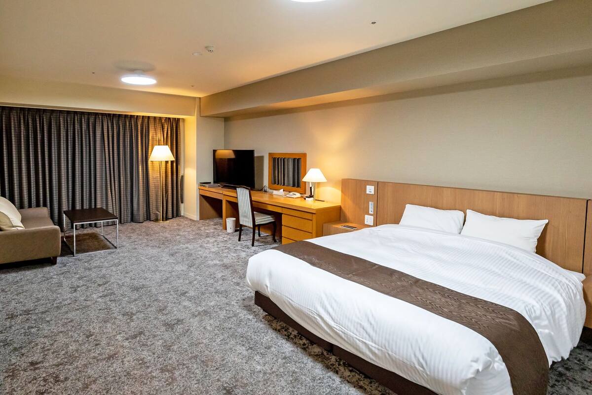 Quarto de hotel com cama, escrivaninha, cadeira, televisão e sofá.
