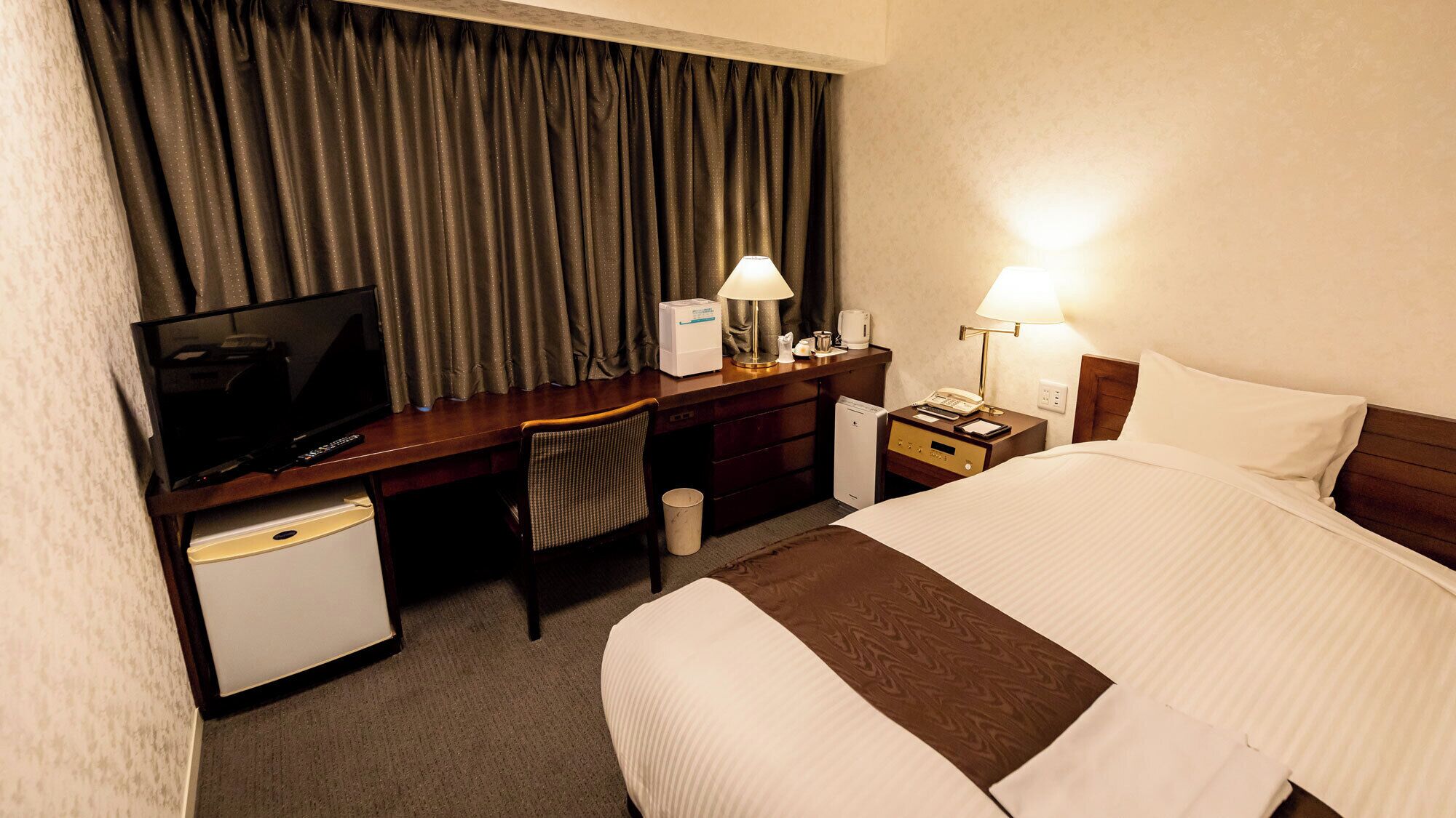Een hotelkamer met een bed, een bureau met televisie, een stoel, een nachtkastje met lamp en een kleine koelkast.