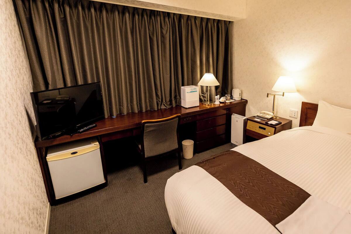 Quarto de hotel com uma cama, uma mesa com televisão, uma cadeira, mesa de cabeceira com luminária e um pequeno frigorífico.