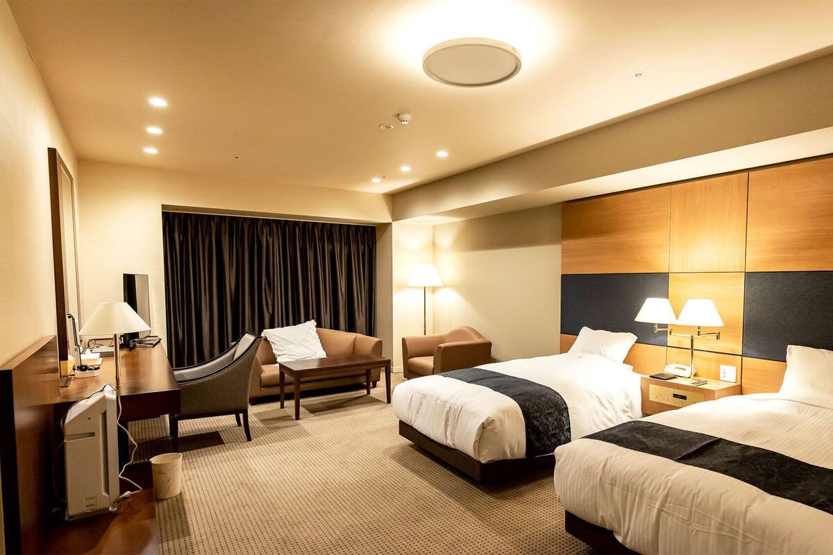 Quarto de hotel com duas camas, uma escrivaninha, uma cadeira, uma luminária e uma janela grande com cortinas.