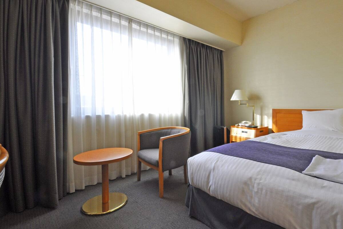 Quarto de hotel com cama, cadeira, mesinha, mesa de cabeceira e abajur.