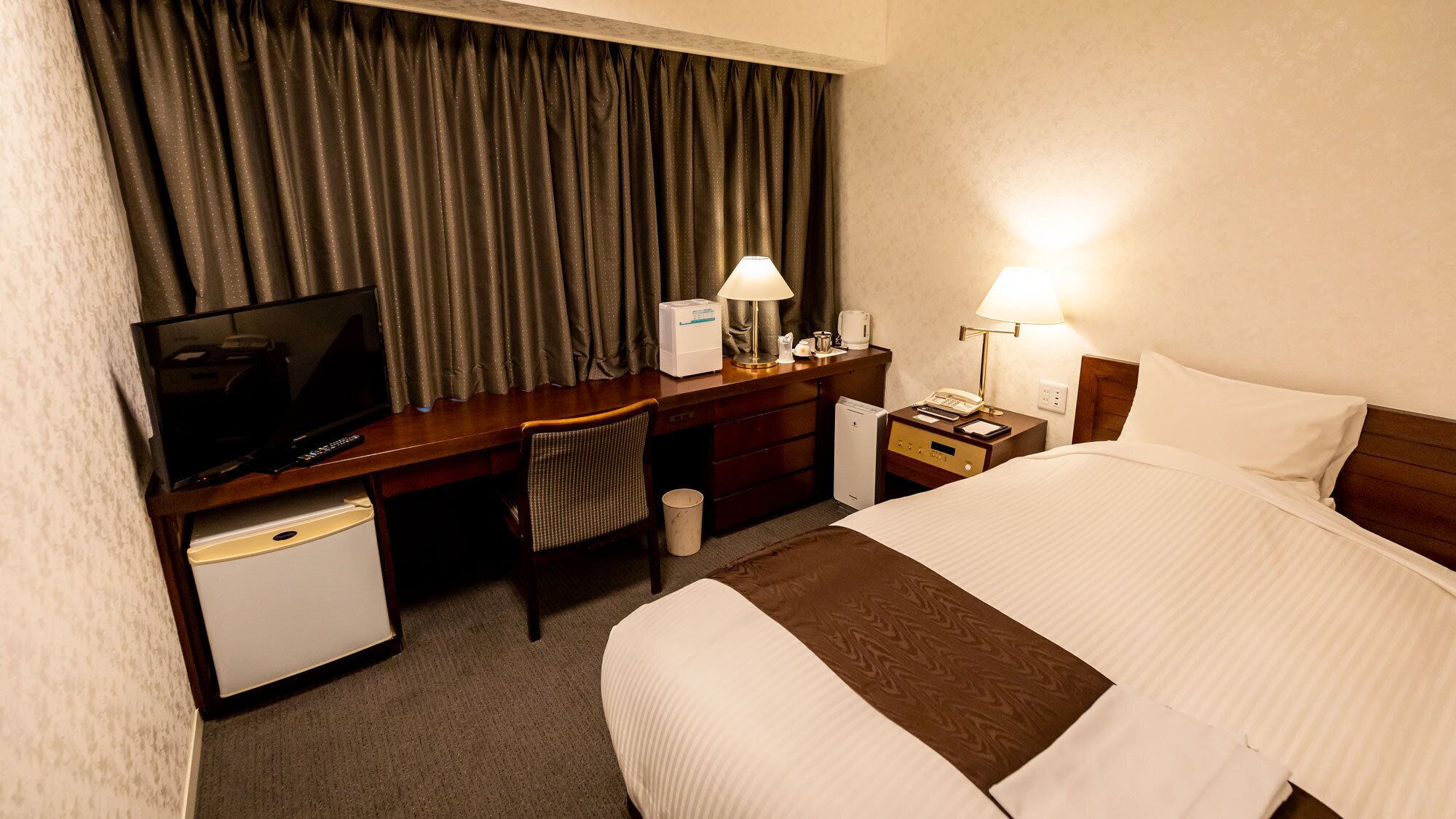 A hotel room with a bed, a desk with a TV, a chair, a nightstand with a lamp, and a small refrigerator.
