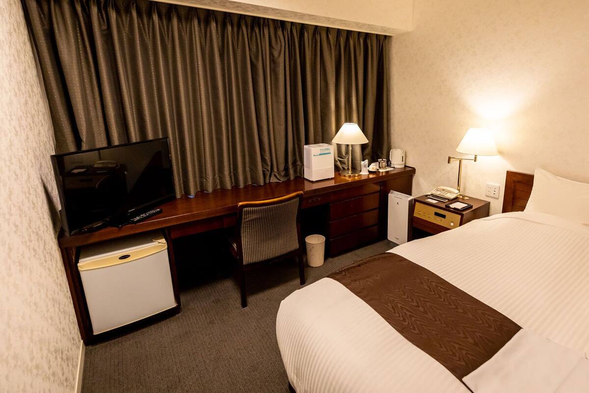 Quarto de hotel com cama, escrivaninha com televisão, cadeira, mesa de cabeceira com abajur e um pequeno frigorífico.