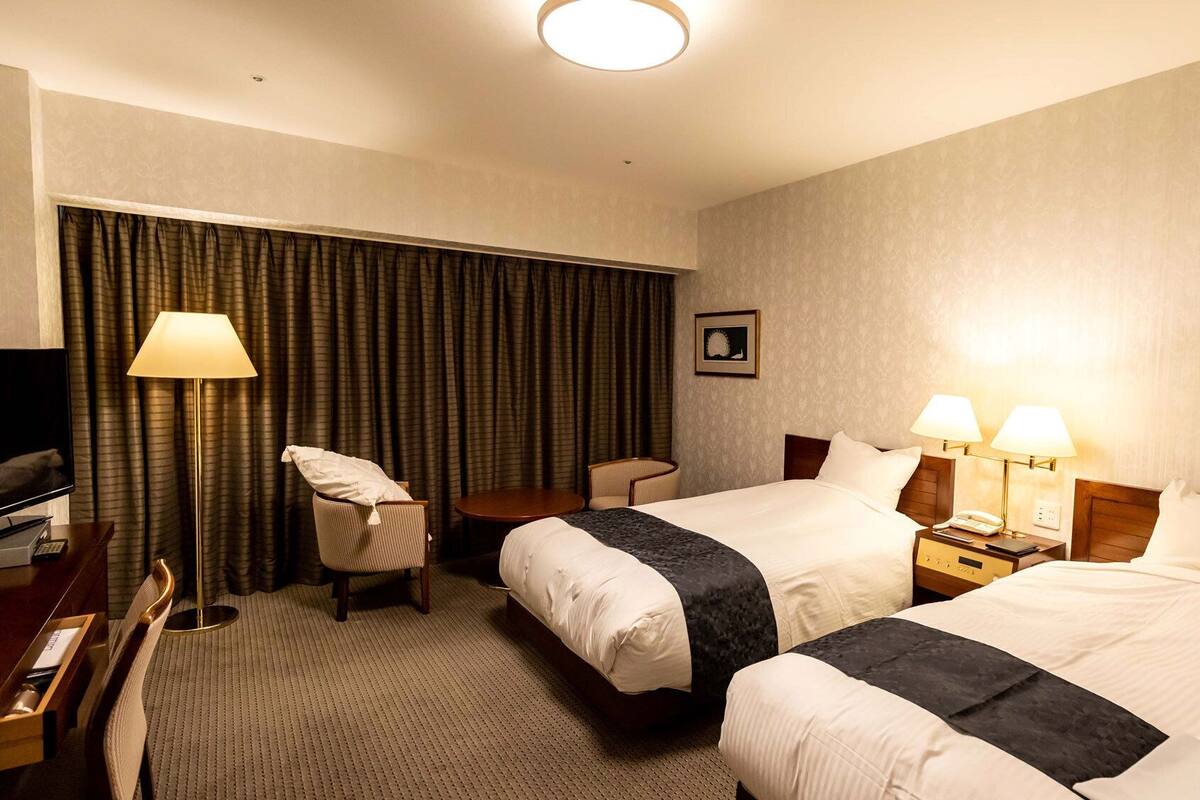 Quarto de hotel com duas camas, uma escrivaninha com televisão, uma cadeira, um abajur e uma mesa de cabeceira com telefone.