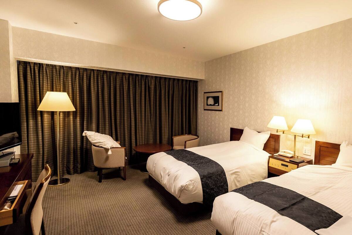 Quarto de hotel com duas camas, uma mesa, uma cadeira, uma televisão e um abajur.