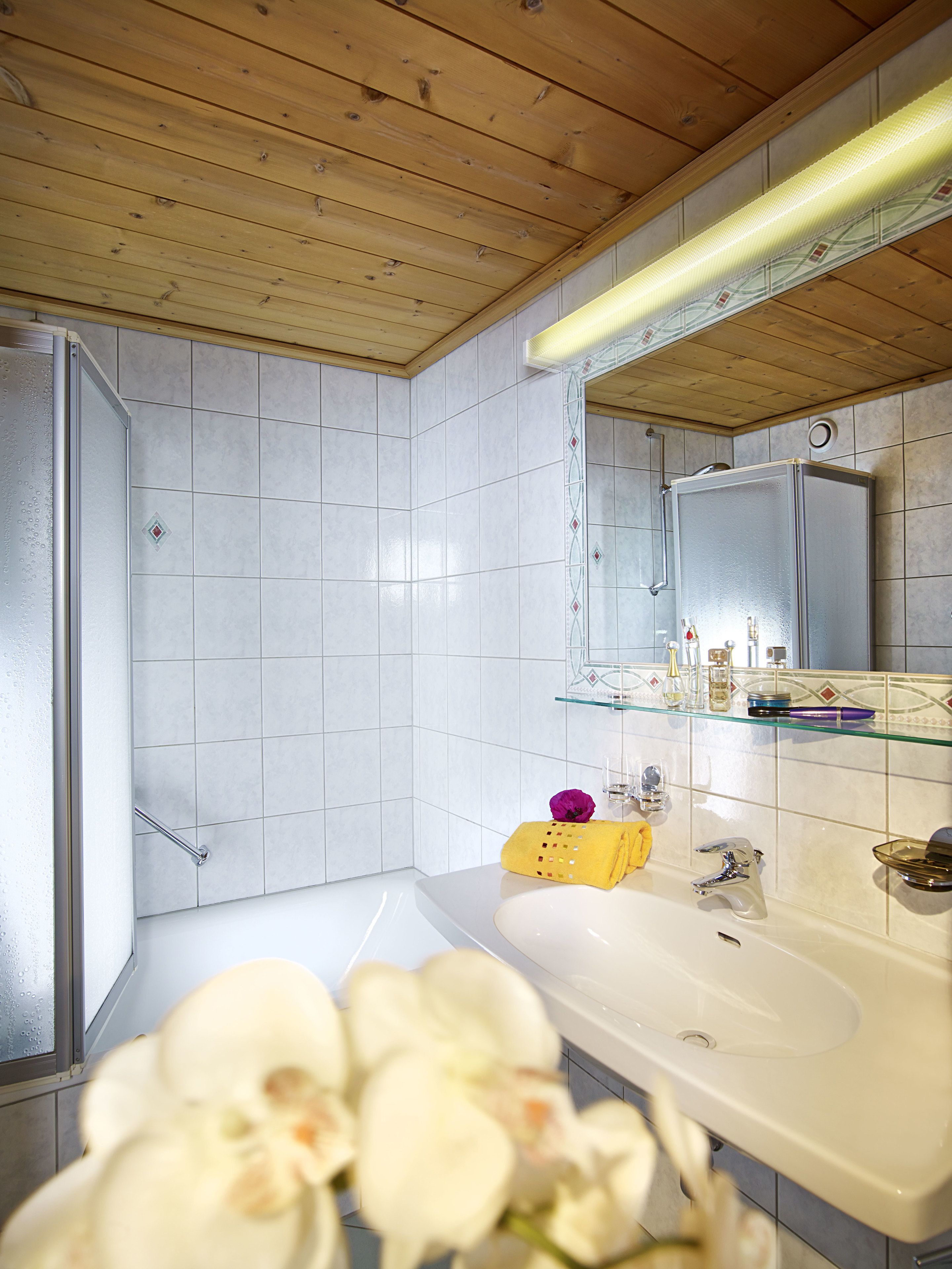 A modern bathroom with a large mirror, white tiles, a wooden ceiling, and a glass shelf with toiletries.