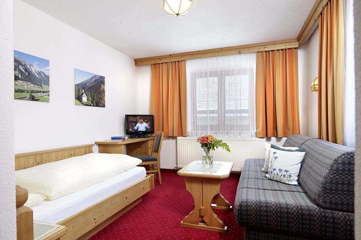 A hotel room with a bed, a sofa, a TV, a small table with a vase of flowers, and two framed pictures on the wall.