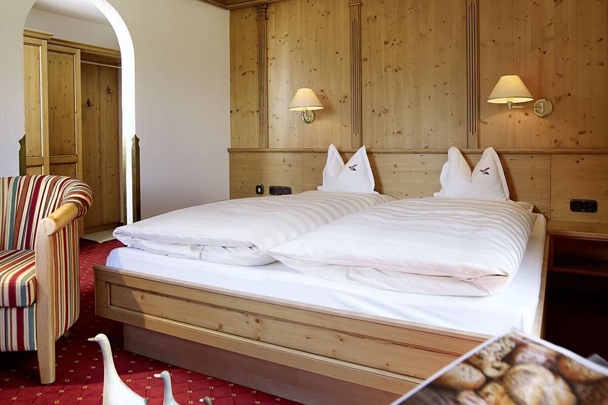 A hotel room with a large bed, wooden headboard, two wall-mounted lamps, a red carpet, a striped armchair, and a door with a wooden frame.