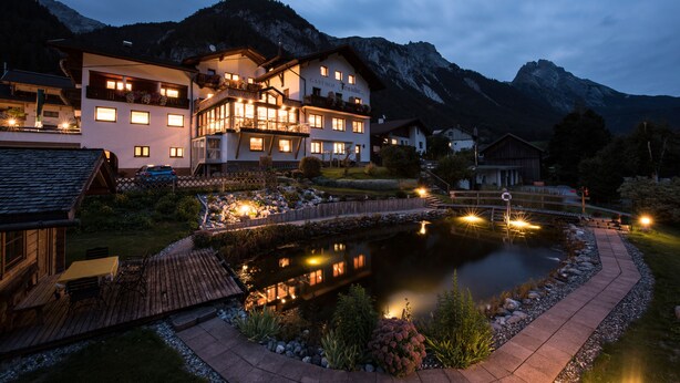 A hotel with a large building, a pond, and mountains in the background. - Hotel Traube (Pettneu am Arlberg)