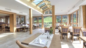 A spacious dining area with wooden tables and chairs, large windows offering a view of the outdoors, and a high ceiling with skylights. - Hotel Traube (Pettneu am Arlberg)
