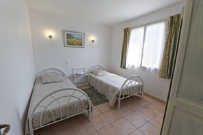 Een slaapkamer met twee eenpersoonsbedden, een klein nachtkastje, een wandlamp, een foto in een lijst en een raam met gordijnen.