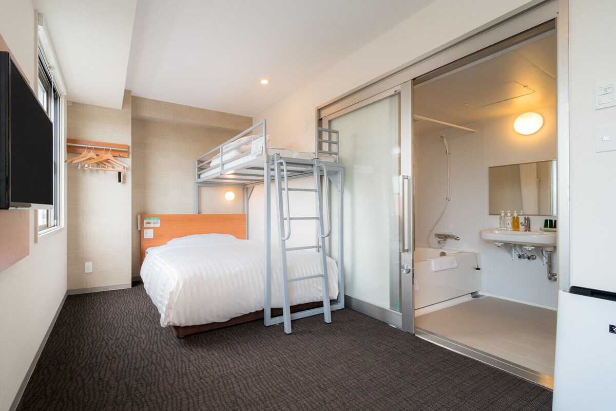 A hotel room with a bunk bed, a flat-screen TV mounted on the wall, a bathroom with a sink and mirror, and a carpeted floor.
