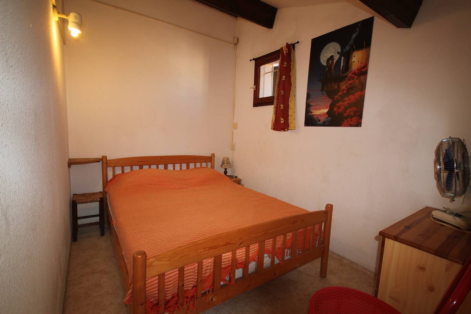 Ein Schlafzimmer mit einem hölzernen Bett, einem Nachttisch, einem Sessel, einem Schreibtisch, einem Ventilator und zwei Bildern an der Wand.