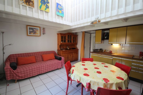 A dining area with a round table covered in a floral tablecloth, surrounded by red chairs. There is a wooden cabinet with glass doors and a red sofa with orange cushions. The room has white walls and a tiled floor.