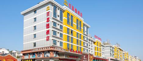 Un hotel de varios pisos con una fachada llamativa en amarillo y blanco, varias tiendas a nivel de la calle y un cielo azul despejado.