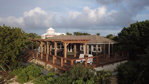 Eine Holzterrasse mit Sitzgelegenheiten im Freien, einem überdachten Bereich und Blick auf ein großes Gebäude im Hintergrund.