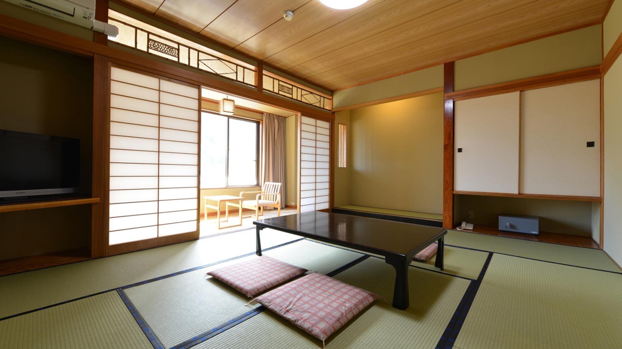 Una habitación japonesa tradicional con suelo de tatami, una mesa baja, cojines, un televisor, puertas correderas y vistas al exterior.