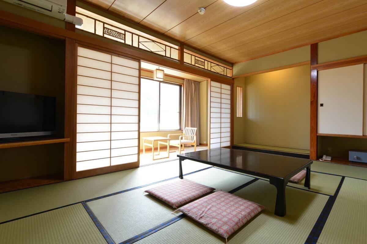 Um quarto japonês tradicional com tatames, uma mesa baixa, almofadas, uma televisão, portas deslizantes e vista para o exterior.